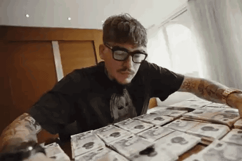 A Man Wearing Glasses Is Sitting At A Table Covered In Money . GIF