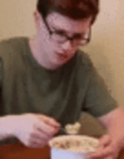 A Man Wearing Glasses Is Sitting At A Table Eating A Bowl Of Food GIF