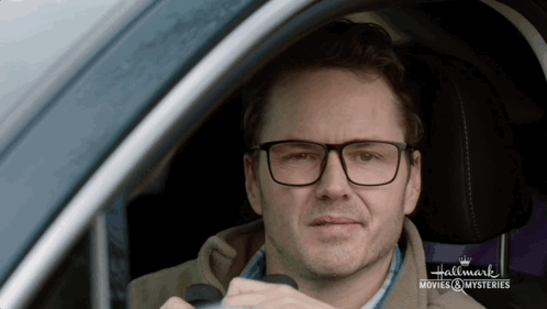 A Man Wearing Glasses Is Sitting In A Car With The Words Hallmark Movies & Mysteries On The Bottom GIF