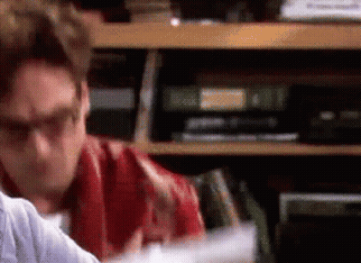 A Man Wearing Glasses Is Sitting In Front Of A Bookshelf Filled With Books GIF