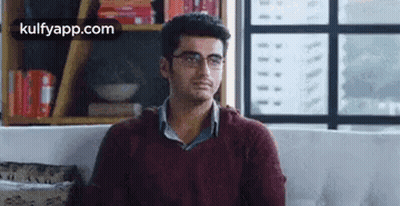 A Man Wearing Glasses Is Sitting On A Couch In A Living Room . GIF