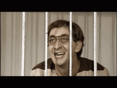 A Man Wearing Glasses Is Smiling Behind Prison Bars GIF