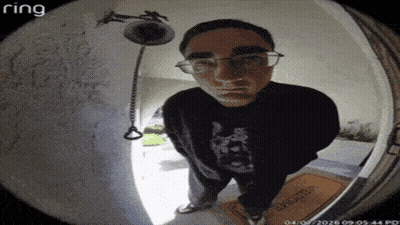 A Man Wearing Glasses Is Standing In Front Of A Doorbell Looking At The Camera . GIF