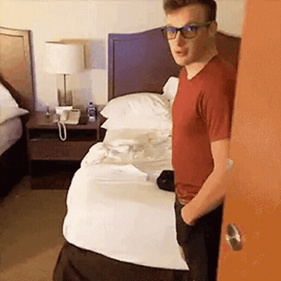 A Man Wearing Glasses Is Standing In Front Of A Hotel Bed . GIF