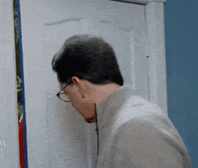 A Man Wearing Glasses Looks Through A Hole In A Door GIF