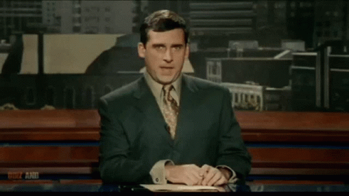 A Man Wearing Glasses Sits At A Desk In Front Of A Sign That Says Ocnv Aaoi GIF