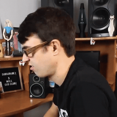 A Man Wearing Glasses Sits In Front Of A Sign That Says " Subscribe 3 Mill Sub Club " GIF
