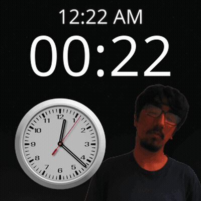 A Man Wearing Glasses Stands In Front Of A Clock That Says 12:22 Am GIF