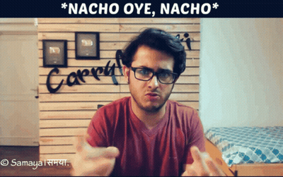 A Man Wearing Glasses Stands In Front Of A Wall That Says Nacho Oye Nacho GIF