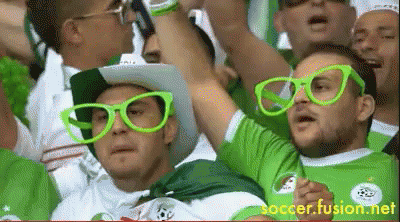 A Man Wearing Green Glasses And A Cowboy Hat Stands In A Crowd Of People GIF