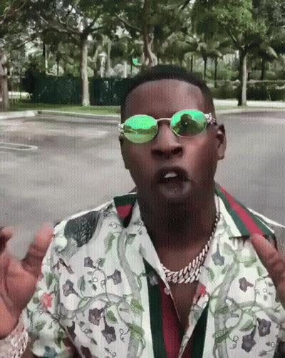 A Man Wearing Green Sunglasses And A Floral Shirt Stands In A Parking Lot GIF