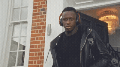 A Man Wearing Headphones And A Black Leather Jacket Stands In Front Of A Brick Building GIF