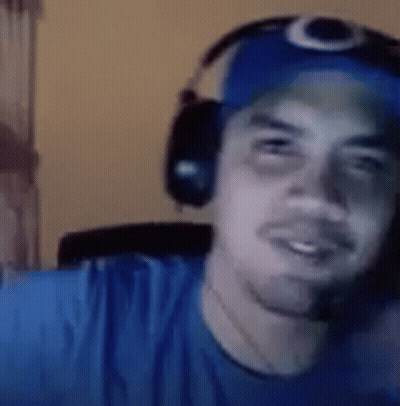 A Man Wearing Headphones And A Blue Hat Is Looking At The Camera . GIF