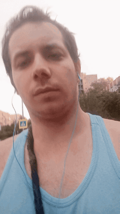 A Man Wearing Headphones And A Blue Tank Top Is Standing In Front Of A Crosswalk Sign GIF