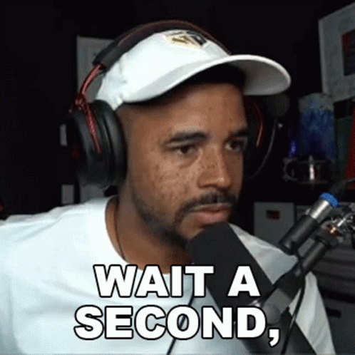 A Man Wearing Headphones And A Hat Is Talking Into A Microphone And Says Wait A Second GIF