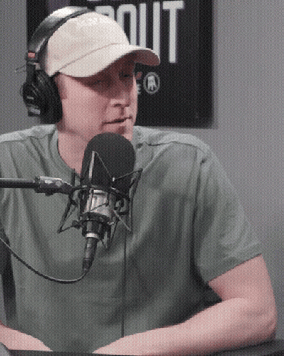 A Man Wearing Headphones And A Hat Talks Into A Microphone In Front Of A Sign That Says Out GIF