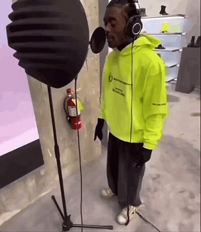 A Man Wearing Headphones And A Neon Yellow Hoodie Sings Into A Microphone GIF