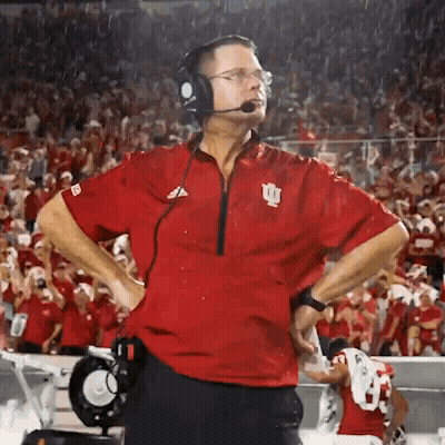 A Man Wearing Headphones And A Red Shirt With The Letter I On It Stands On A Football Field . GIF