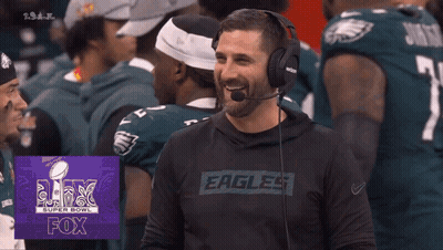 A Man Wearing Headphones And A Shirt That Says Eagles Laughs In Front Of Football Players GIF
