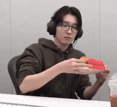 A Man Wearing Headphones And Glasses Is Sitting At A Table Eating A Sandwich . GIF