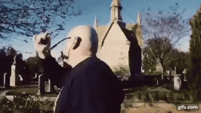 A Man Wearing Headphones And Glasses Is Standing In Front Of A Church In A Cemetery . GIF