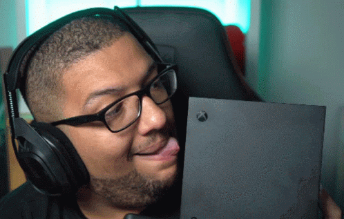 A Man Wearing Headphones And Glasses Is Sticking His Tongue Out At An Xbox Box GIF