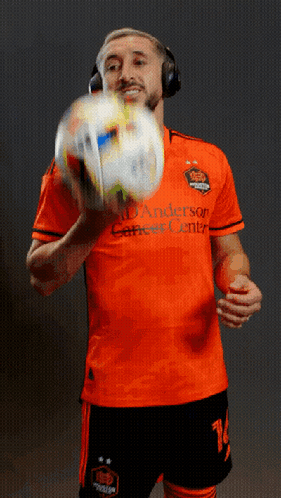 A Man Wearing Headphones Holds A Soccer Ball While Wearing An Orange Anderson Career Center Jersey GIF