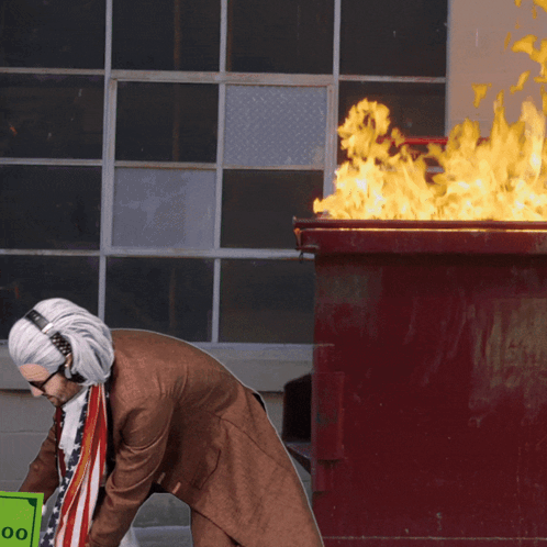 A Man Wearing Headphones Is Holding A 100 Dollar Bill In Front Of A Dumpster On Fire GIF