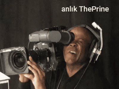 A Man Wearing Headphones Is Holding A Camera And Smiling In Front Of A Sign That Says Anlik Theprine GIF