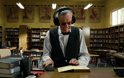 A Man Wearing Headphones Is Reading A Book In A Library With Posters Of Darwin And Albert Einstein On The Wall GIF