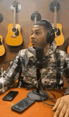 A Man Wearing Headphones Is Sitting At A Table In Front Of A Microphone In Front Of A Wall Of Guitars . GIF