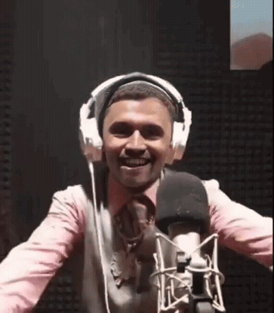 A Man Wearing Headphones Is Standing In Front Of A Microphone And Smiling . GIF