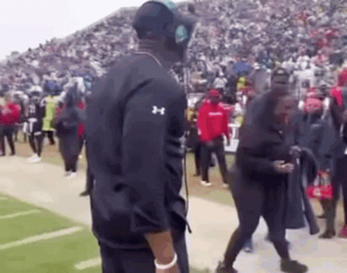 A Man Wearing Headphones Is Standing On A Football Field With A Crowd Of People . GIF