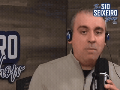 A Man Wearing Headphones Is Talking Into A Microphone In Front Of A Sign That Says The Sid Seixeiro Show GIF