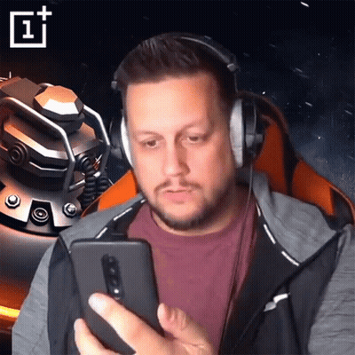 A Man Wearing Headphones Looks At His Phone With A Oneplus Logo Behind Him GIF