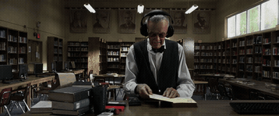 A Man Wearing Headphones Sits At A Desk In A Library Reading A Book GIF