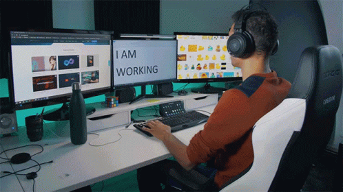 A Man Wearing Headphones Sits At A Desk With Three Computer Monitors And The Words I Am Working On The Screen GIF