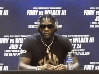 A Man Wearing Headphones Sits At A Table With A Bottle Of Water In Front Of A Sign That Says Fury GIF