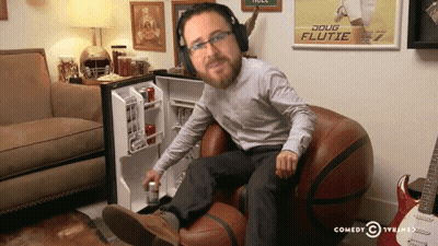 A Man Wearing Headphones Sits In A Chair In Front Of A Doug Flutie Poster GIF