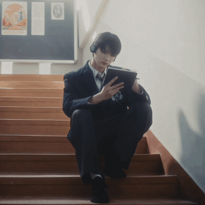 A Man Wearing Headphones Sits On A Set Of Stairs Looking At His Phone GIF