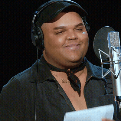 A Man Wearing Headphones Stands In Front Of A Microphone With A Necklace That Says Exi GIF