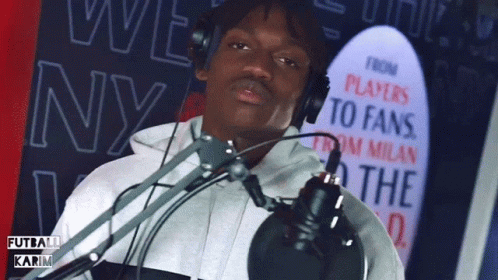A Man Wearing Headphones Stands In Front Of A Microphone With A Sign Behind Him That Says " Players To Fans " GIF