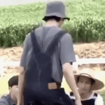 A Man Wearing Overalls And A Hat Is Standing In A Field With A Backpack . GIF