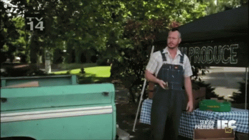 A Man Wearing Overalls Stands In Front Of A Produce Stand GIF