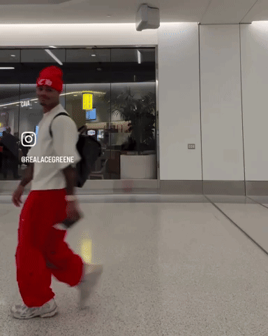 A Man Wearing Red Pants And A Red Hat Is Walking In A Hallway With A Sign That Says @Realacegreene GIF