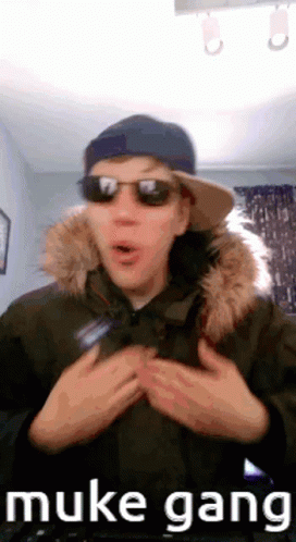 A Man Wearing Sunglasses , A Hat And A Parka With A Fur Hood Is Standing In A Room . GIF