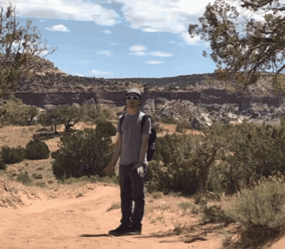 A Man Wearing Sunglasses And A Backpack Stands On A Dirt Road In The Desert GIF