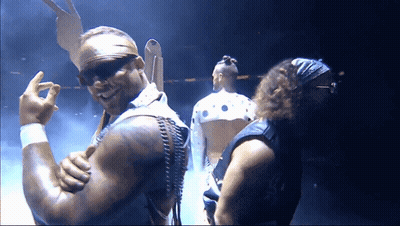 A Man Wearing Sunglasses And A Bandana Stands Next To Another Man In A Dark Room GIF