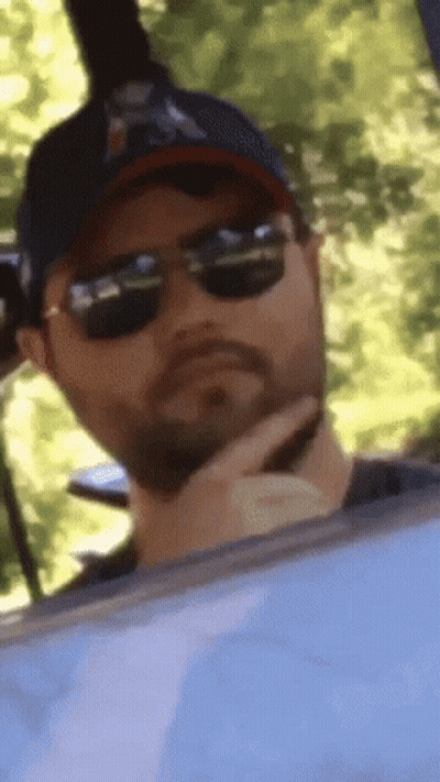 A Man Wearing Sunglasses And A Baseball Cap Is Looking Out Of A Window GIF
