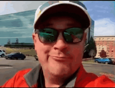 A Man Wearing Sunglasses And A Baseball Cap Is Smiling In Front Of A Building . GIF